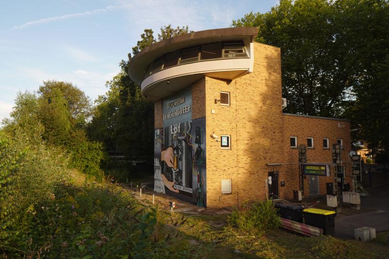 Ein Stellwerksgebäude mit einer halbrunden Kanzel und der Aufschrift ‘Bochum Langendreer Lpf’. Die Vorderseite ist bemalt mit Kabeln, Relaisanlagen und einem Signal. Vor dem Eingang steht ein Abfahrtsanzeiger und einige Signalanlagen.